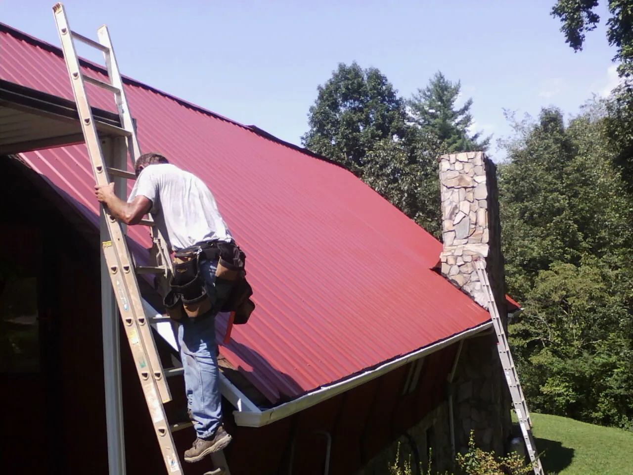 Roofer on a ladder beside a red metal roof and stone chimney — Soffit & Fascia Repair by Roofing Brothers in Port Costa